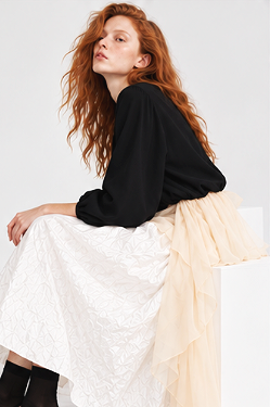 A woman with long, curly red hair wearing a black top and a beige tulle skirt, seated against a plain white background.