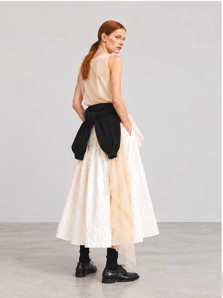 A woman with red hair wearing a sleeveless beige top, a long white textured skirt with a sheer beige overlay, black boots, and a black sweater tied around her waist, standing against a plain gray wall.