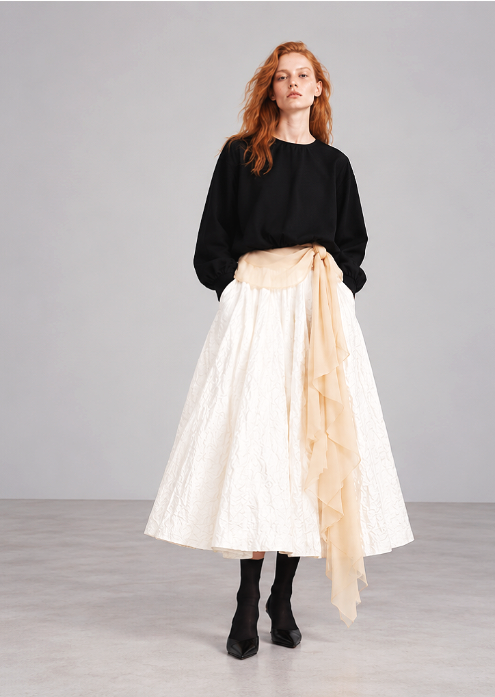 Woman with red hair wearing a black top, white full-length skirt with textured pattern, cream-colored sash with ruffled fabric, black tights, and black shoes, standing against a gray background.