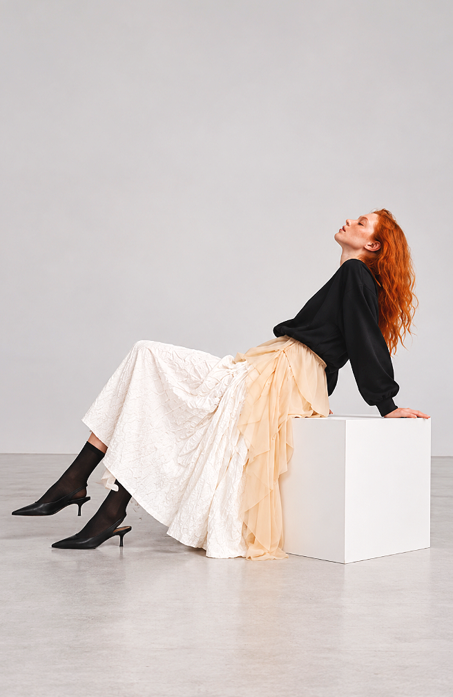 A woman with long red hair wearing a black top, beige layered skirt, black heels, and black sheer socks, sitting on a white block with her head tilted back in a relaxed pose against a plain light gray background.