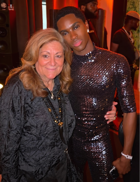 Two women posing together at an indoor event, with a woman on the left having shoulder-length blonde hair and wearing a black textured jacket and layered necklaces, and a woman on the right with short black hair and wearing a shiny black sequined dress.