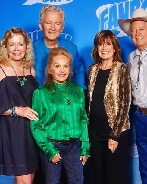 Five older adults, three women and two men, posing together at an event with a blue backdrop. They are smiling and dressed in casual to semi-formal attire, standing close together.