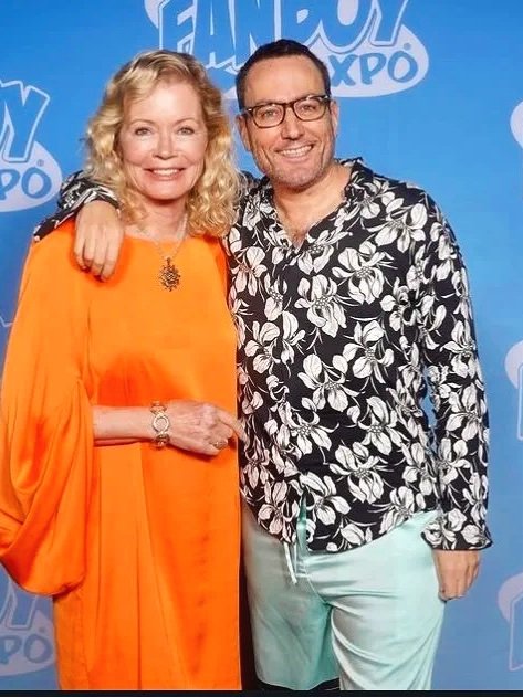 A woman in an orange dress and a man in a black and white floral shirt posing together at Fan Expo, with a blue backdrop featuring the Fan Expo logo.