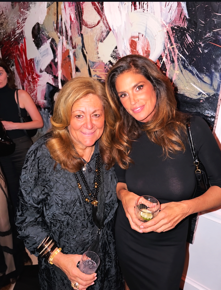 Two women at an art gallery, standing close, smiling, holding drinks, with an abstract painting in the background.