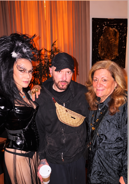 Three people posing together indoors, with orange curtains in the background and a dark artwork on the wall.