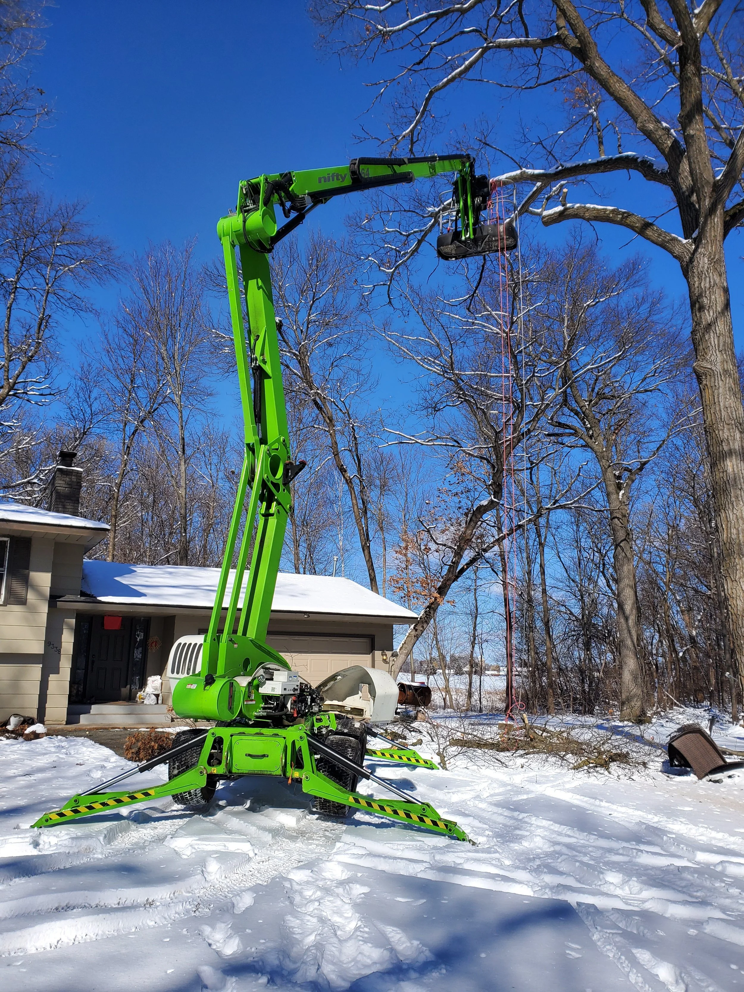 A bright green electric lift tree service crane extends upward around snow-covered trees in a residential yard with blue sky and clear weather.