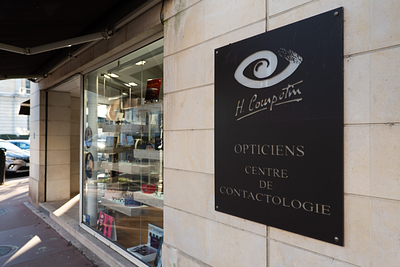 Panneau en métal noir avec le texte blanc : 'H. Couprie, Opticiens, Centre de Contactologie' fixé sur un mur en pierre à l'extérieur d'une boutique d'optique à vitrine en verre.