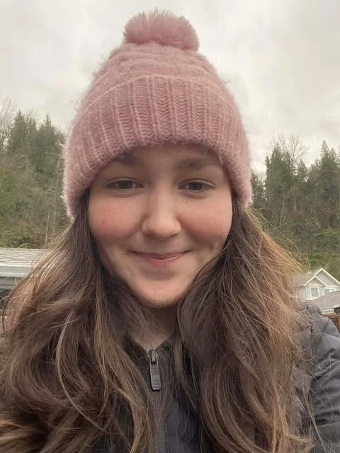 A young woman wearing a pink knit beanie with a pom-pom on top, smiling outdoors with trees and a house in the background.