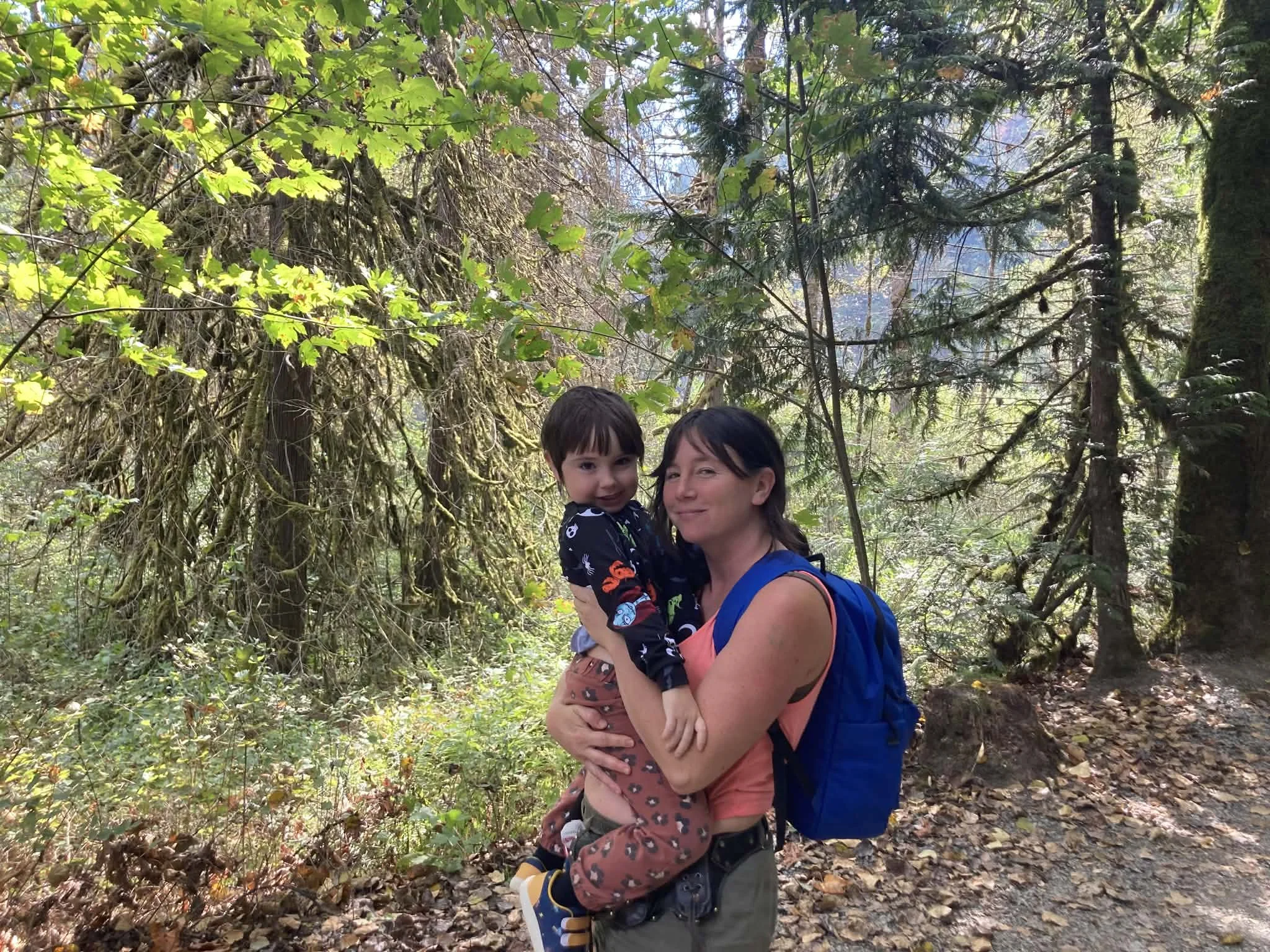 A woman carrying a young boy on her arms while hiking in a forest surrounded by tall trees and lush green foliage.