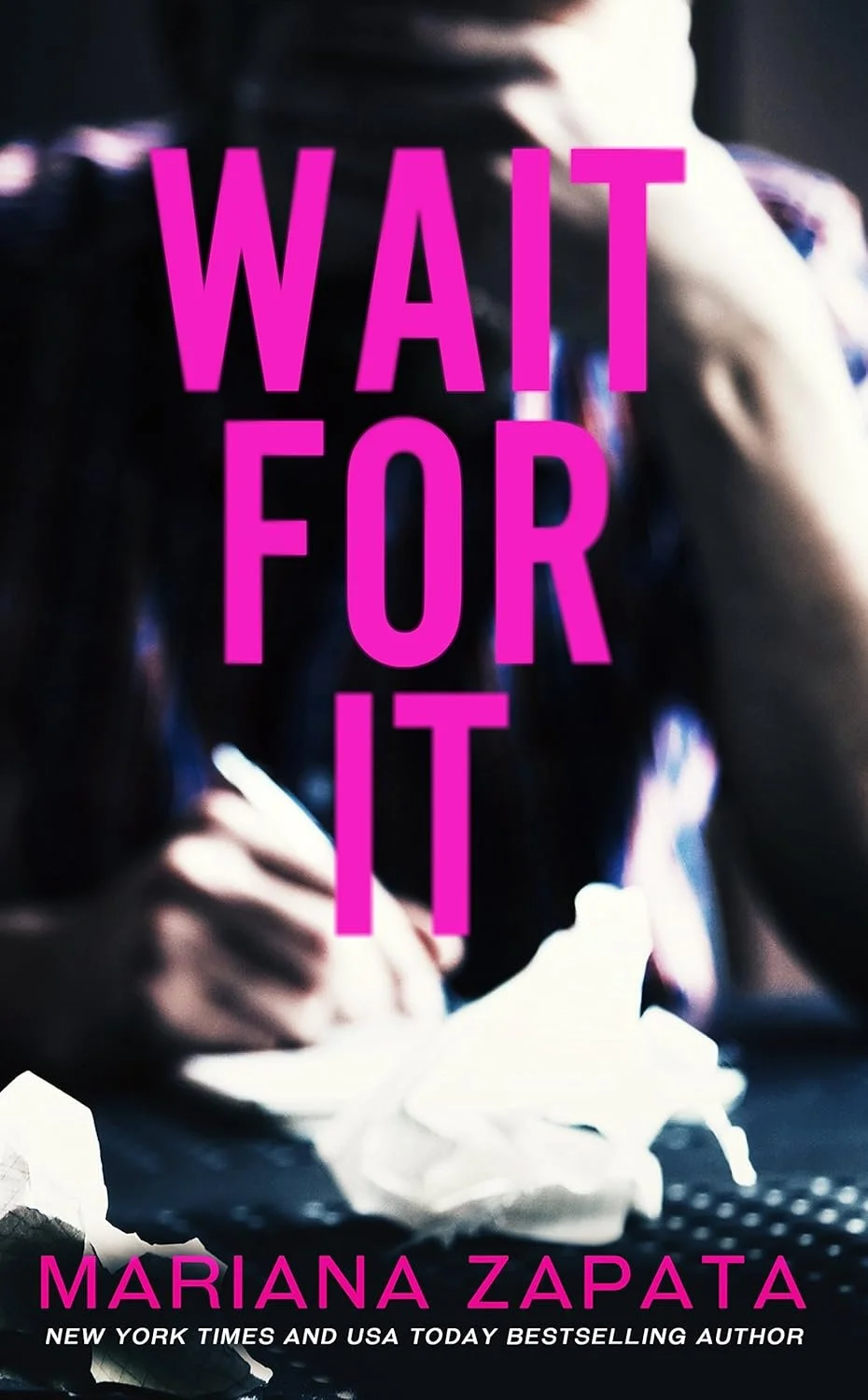 Book cover with the title "Wait For It" in large pink letters, showing a blurry background of a person writing with a pen surrounded by crumpled papers, and author's name Marian Zapata at the bottom.