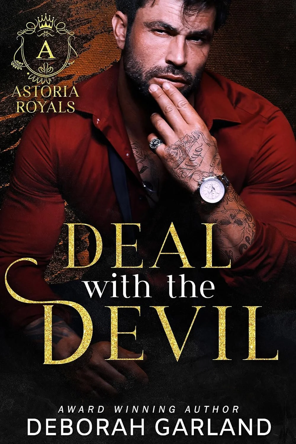 Book cover featuring a man with tattoos and a beard, wearing a red shirt, with a serious expression, hand near his face, and a tattoo on his arm. The title reads 'Deal with the Devil' by Deborah Garland, with gold and white text, and a decorative emblem with a crown and the letter 'A' at the top left corner.