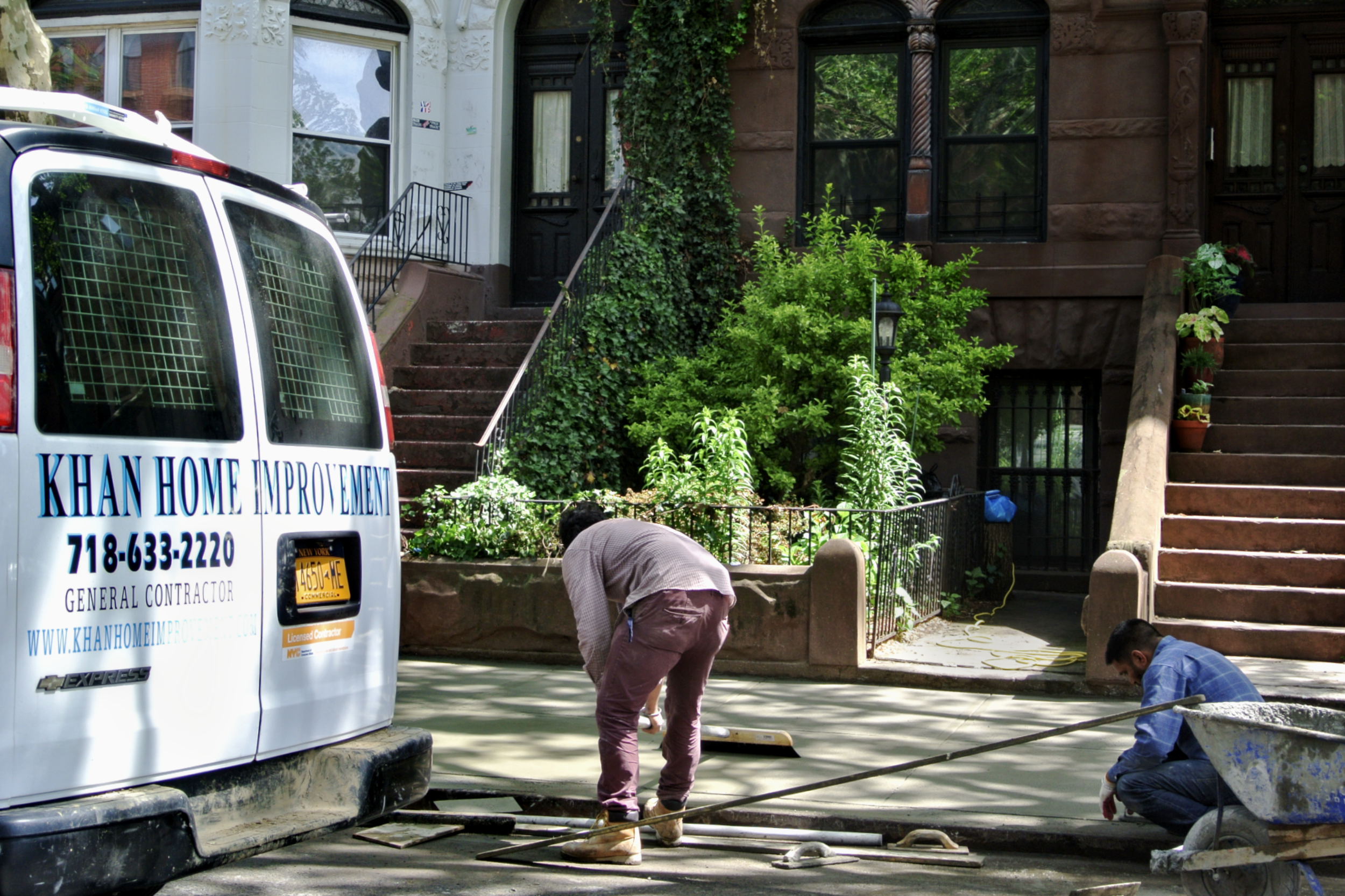 Two masonry contractors are resurfacing a sidewalk in front of residential brownstone buildings with stairs in Brooklyn, next to an official Khan Home Improvement’s van.
