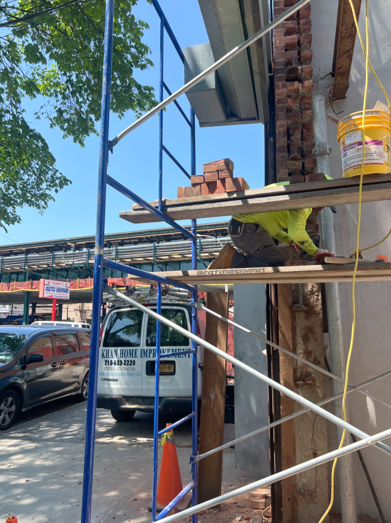 Construction worker crouched down on scaffolding, working on a brick wall exterior of a building on a sunny day. A Khan Home Improvement truck is parked nearby, and there are several cars on the street.