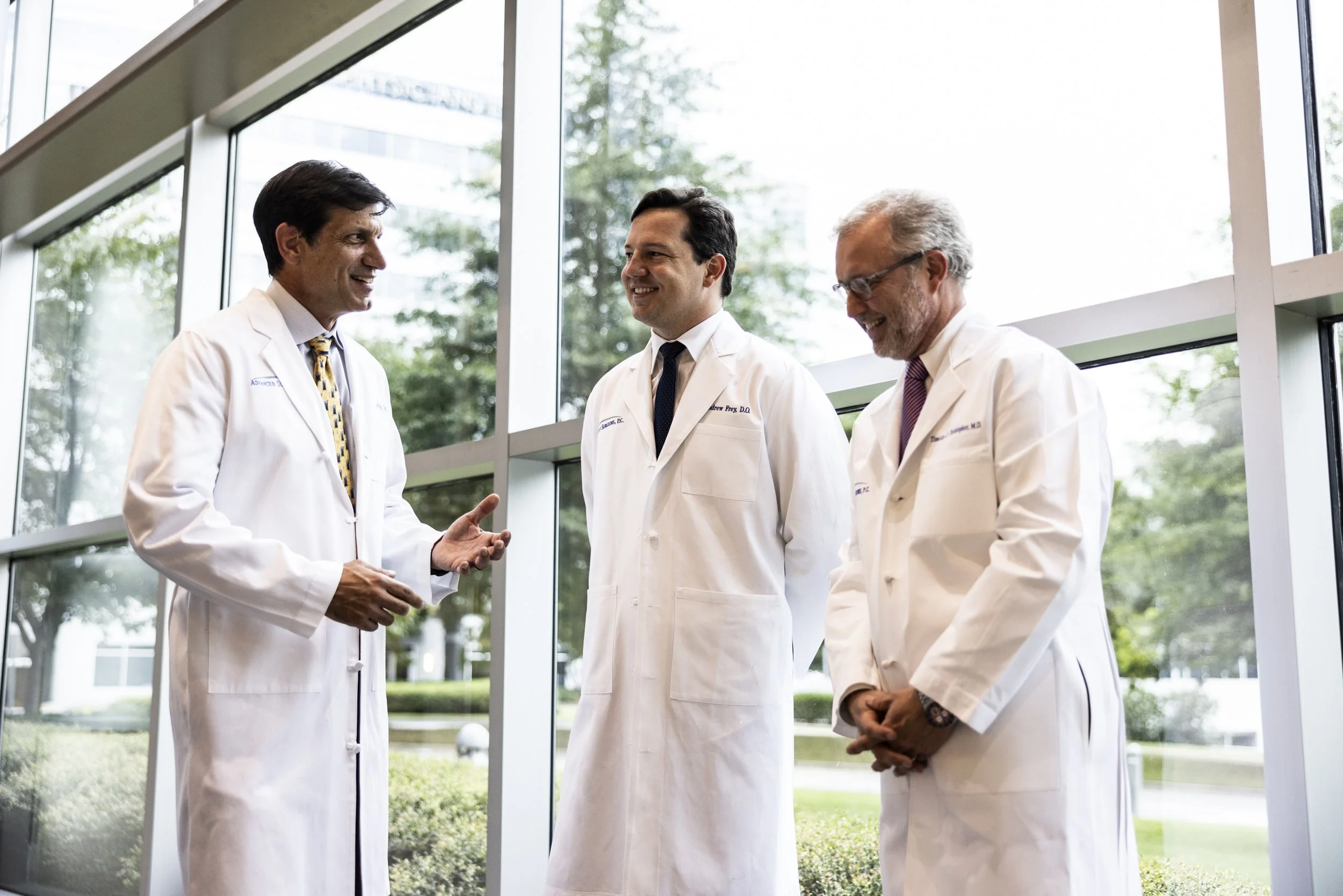 Three doctors in white coats standing and talking in a hospital corridor with large windows.