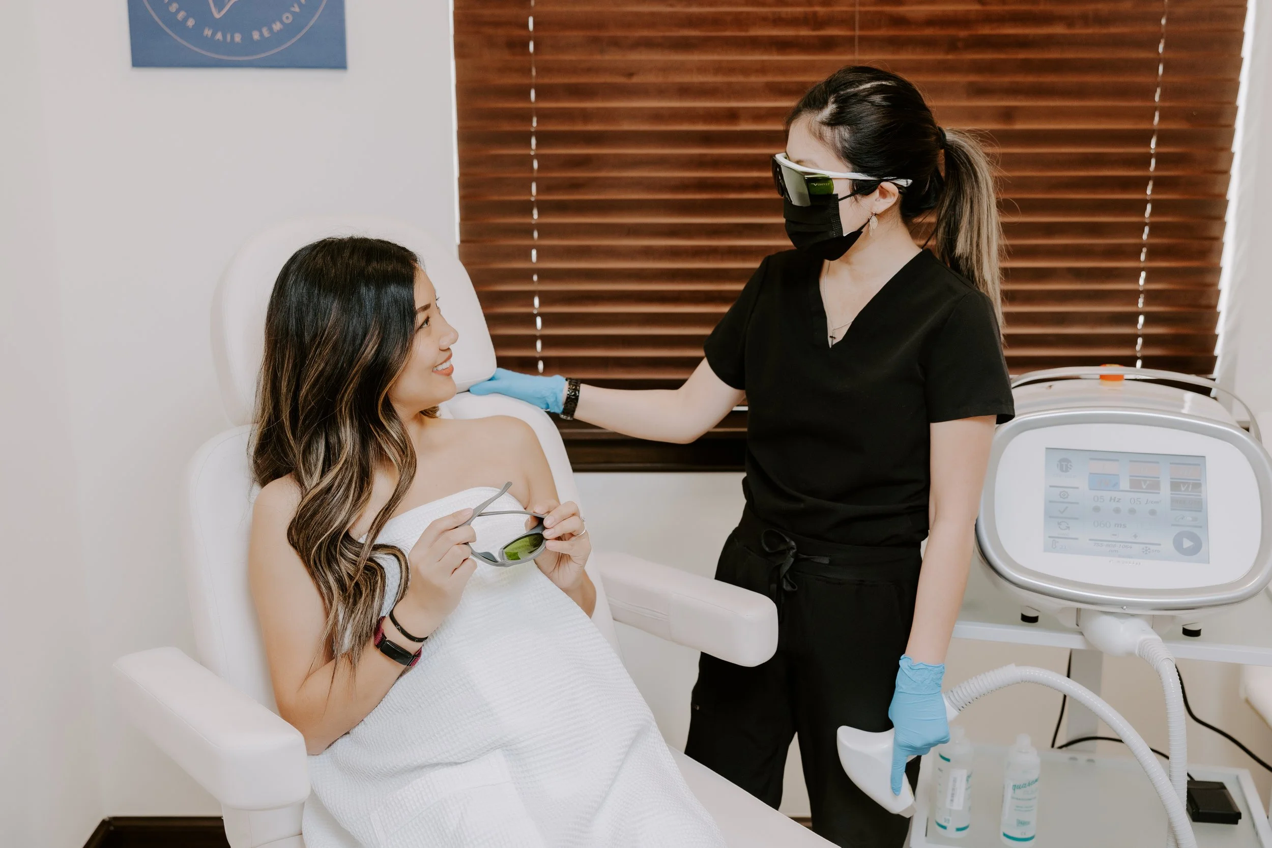 A woman sitting in a medical chair at a clinic, smiling and holding sunglasses, while a healthcare professional in black scrubs and protective gear performs a procedure with medical equipment nearby.
