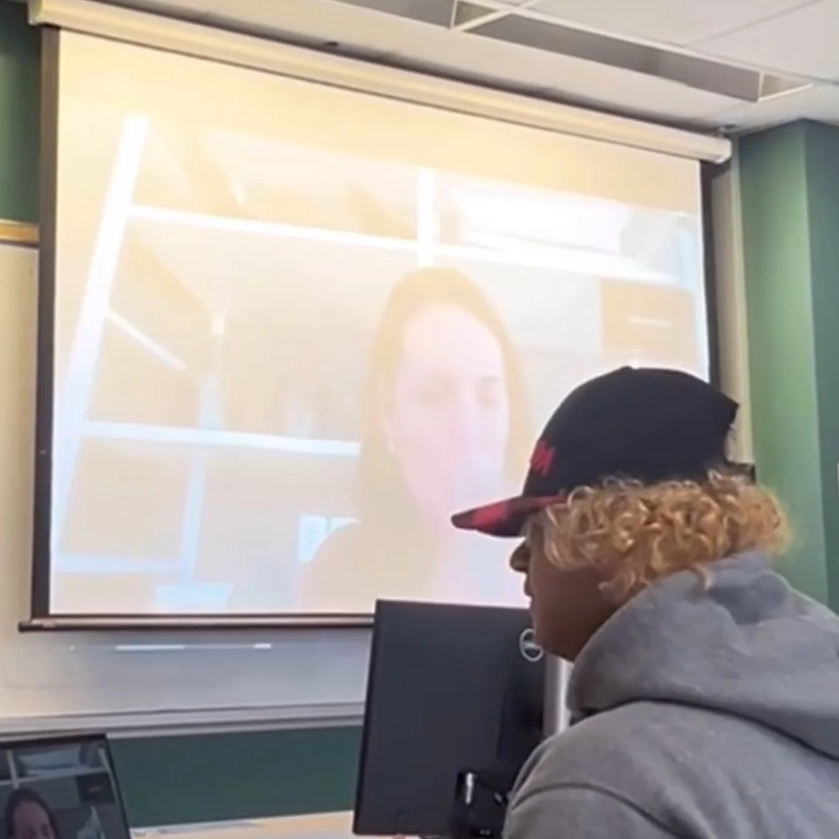 A person in a classroom watching a virtual presentation on a projector screen. A computer monitor and laptop are also visible.