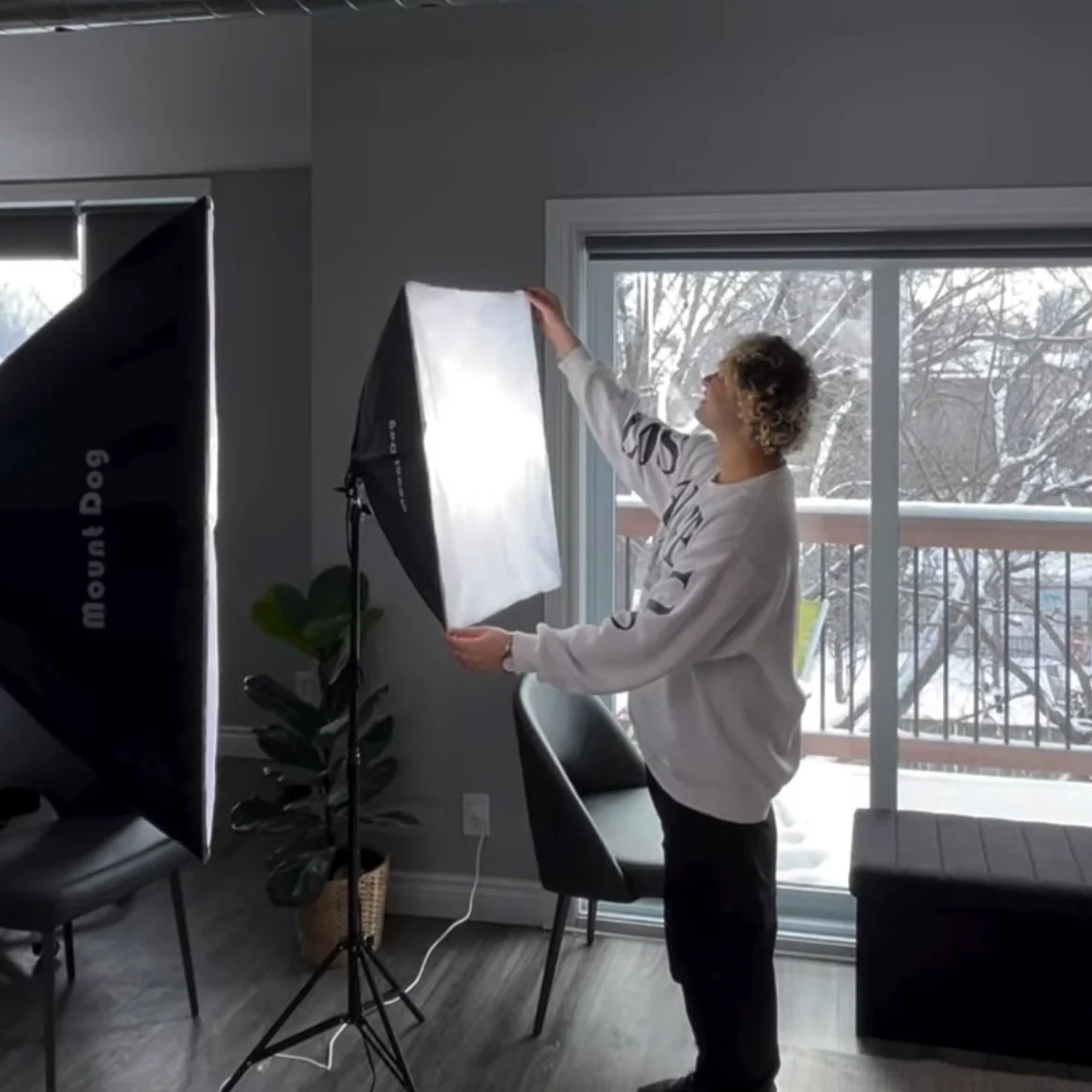 Person adjusting a photography softbox light in a room with snow visible outside the window.