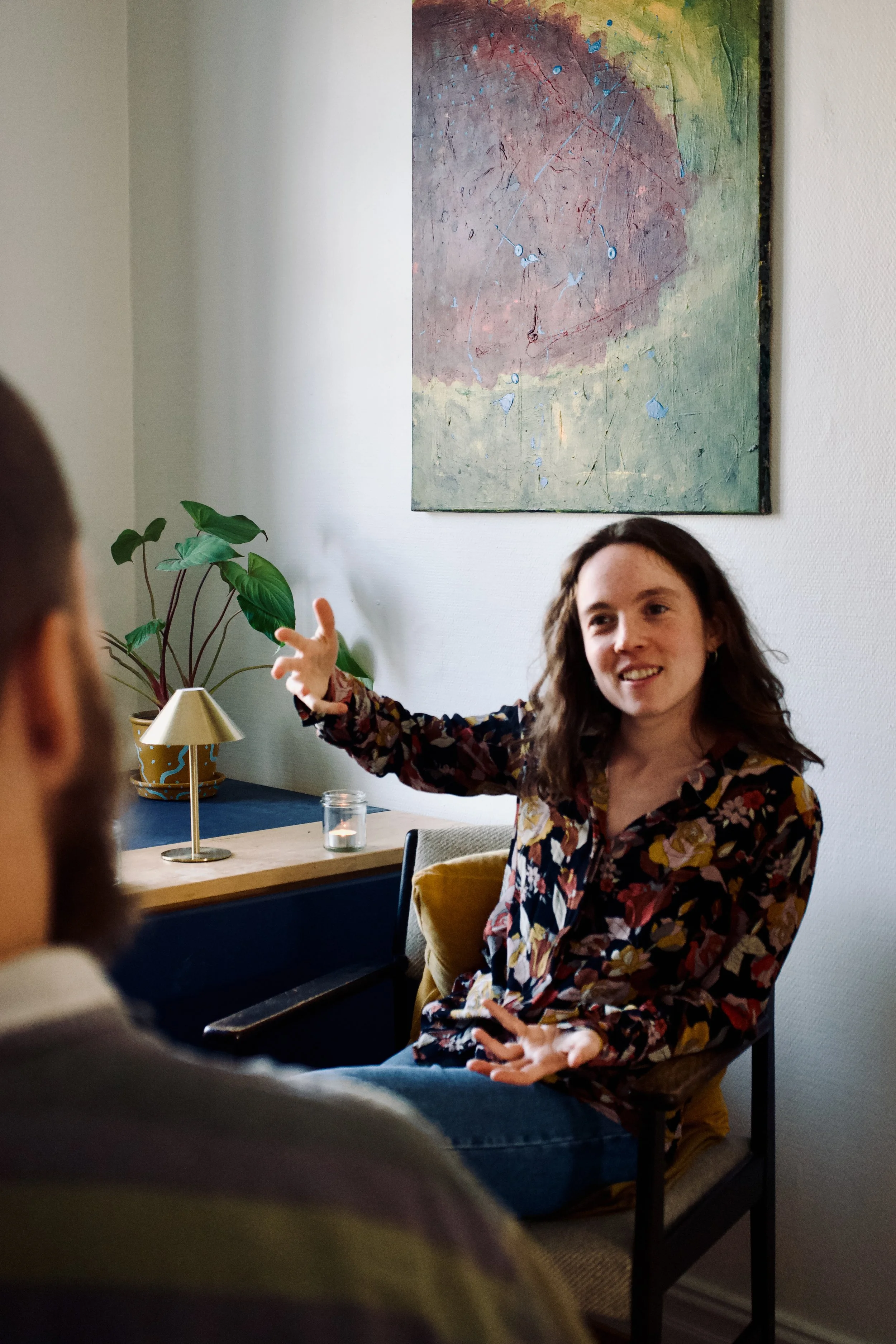 En kvinde sidder og taler under en samtale, og peger med hånden, mens en person i forgrunden lytter. Baggrunden har et abstrakt maleri og et grønt plante på et bord.