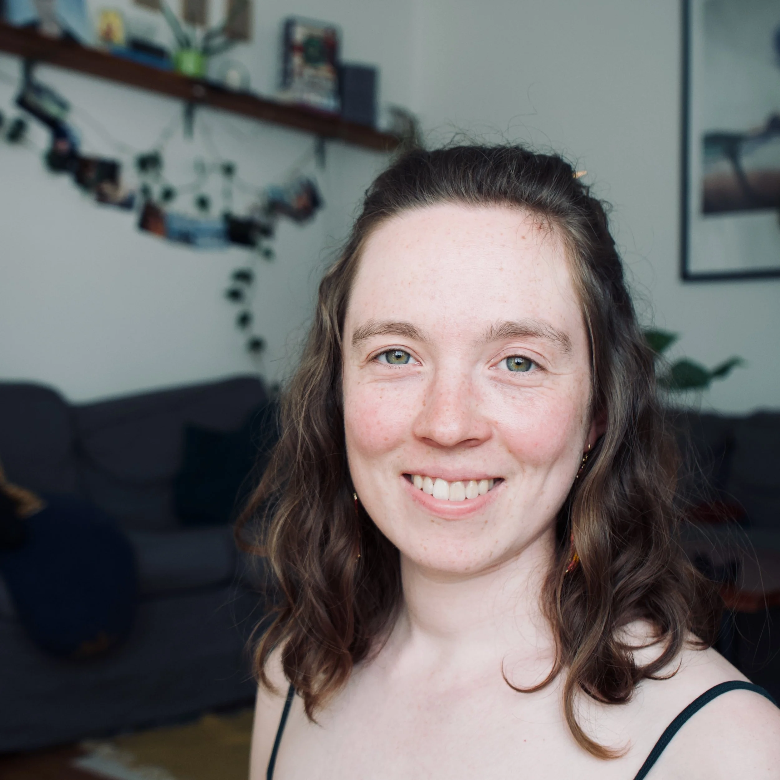 Billedet viser en person med brunt, bølget hår og blå øjne, der smiler. Personens ansigt er centreret i billedet. Baggrunden er en indretning med hylder og billeder på væggen.