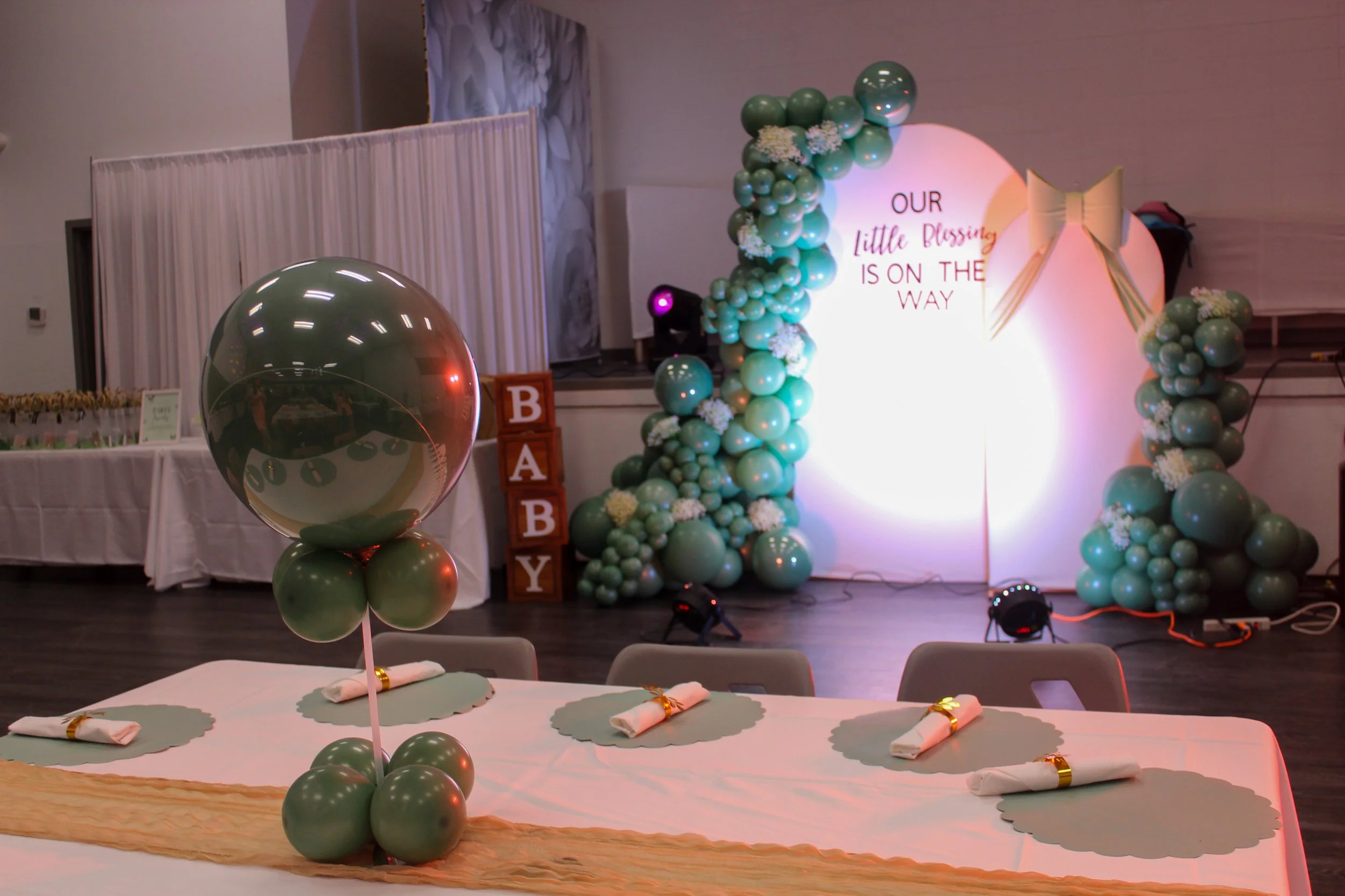 Decorated celebration table with a black balloon and green balloons, set with napkins and gold rings in front of a backdrop with turquoise balloons, white flowers, and a sign that reads "Our little blessing is on the way".