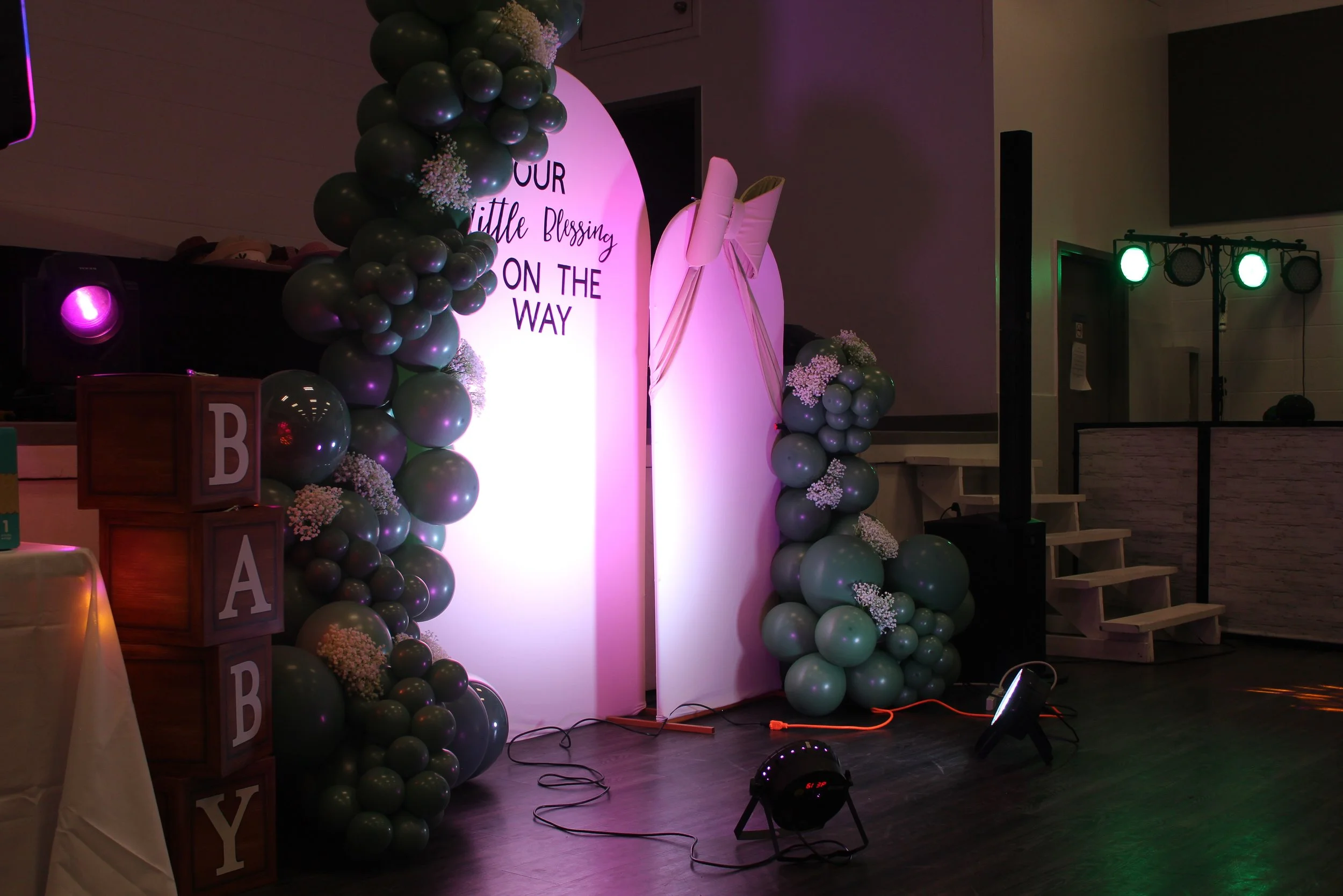 Decorated event space with balloon arch, floral accents, and a backdrop with a message about a blessing on the way, likely for a baby announcement or shower.