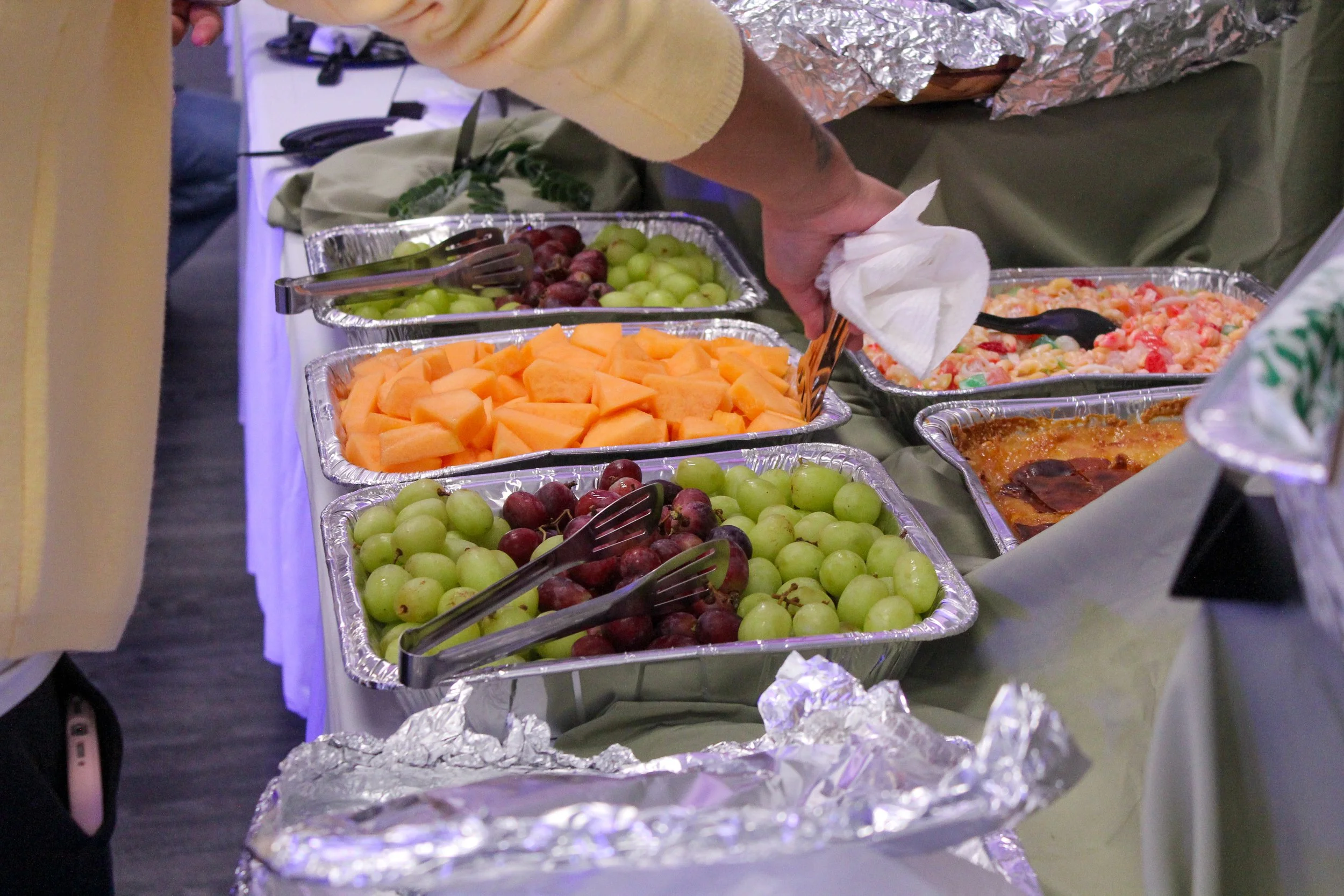 A buffet table with aluminum trays containing cut cantaloupe, green grapes, red grapes, pasta salad, and a dish with sauce. A person is serving themselves using tongs.