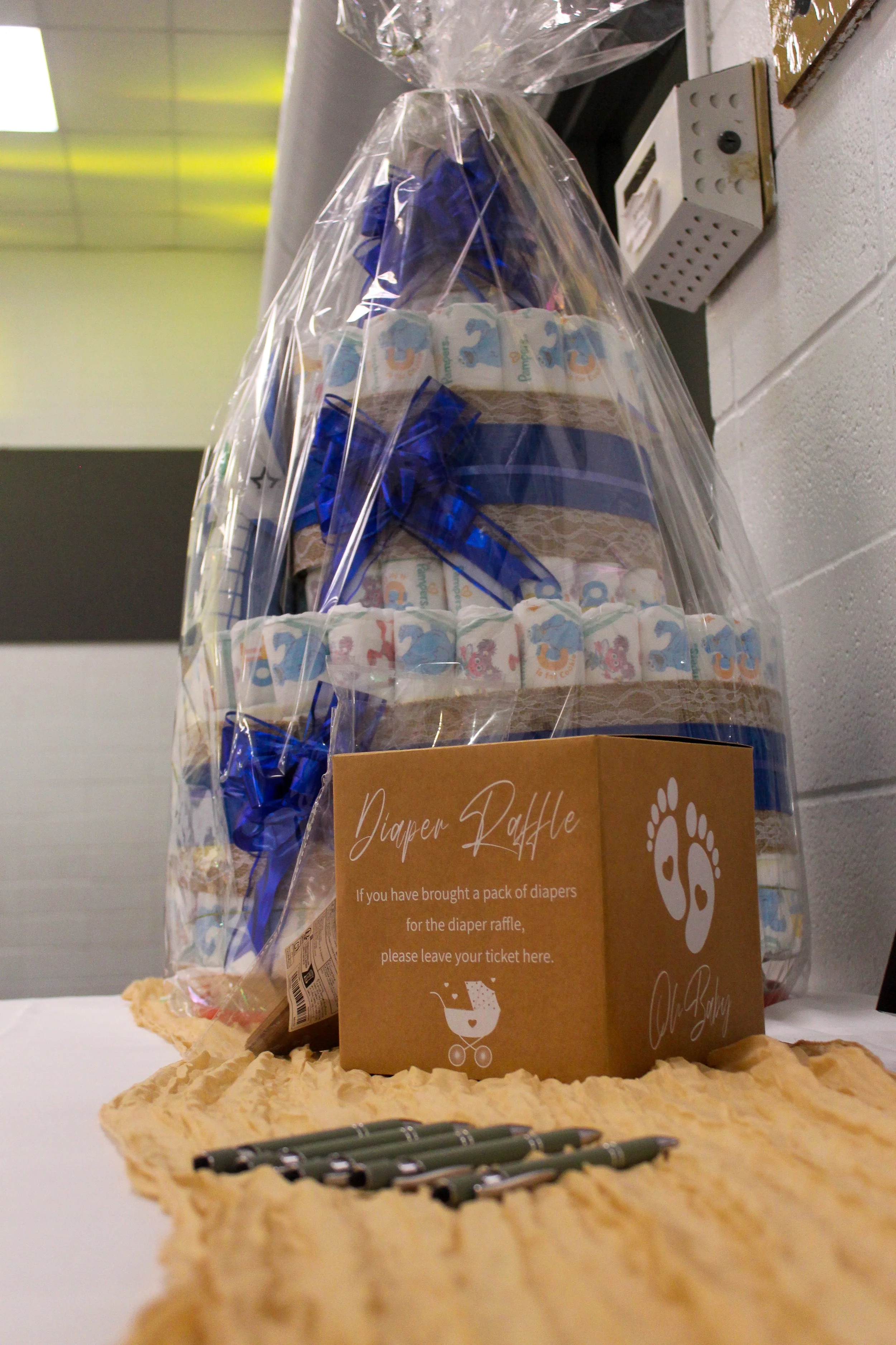 A large diaper raffle basket wrapped in clear plastic, filled with packs of diapers decorated with animal prints, and adorned with a blue ribbon, placed on a table with pens and a tissue paper runner. A sign on the basket reads 'Diaper Raffle' and in
