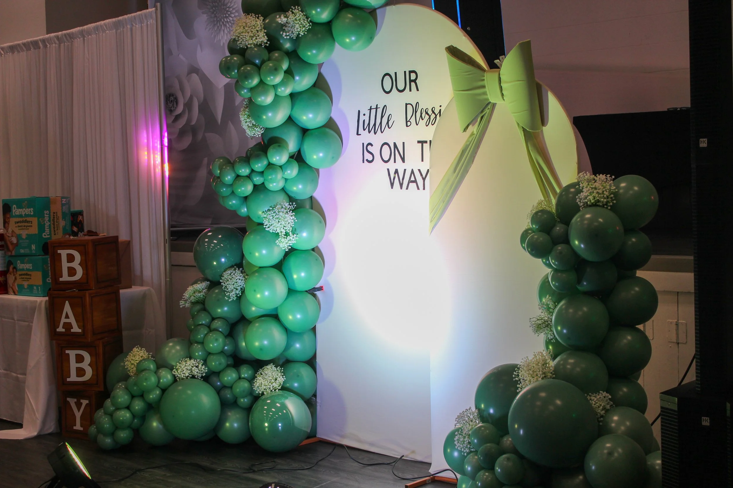 Decorated baby shower arch with green balloons, a large bow, and a sign that reads 'Our little blessing is on the way' in an event space.