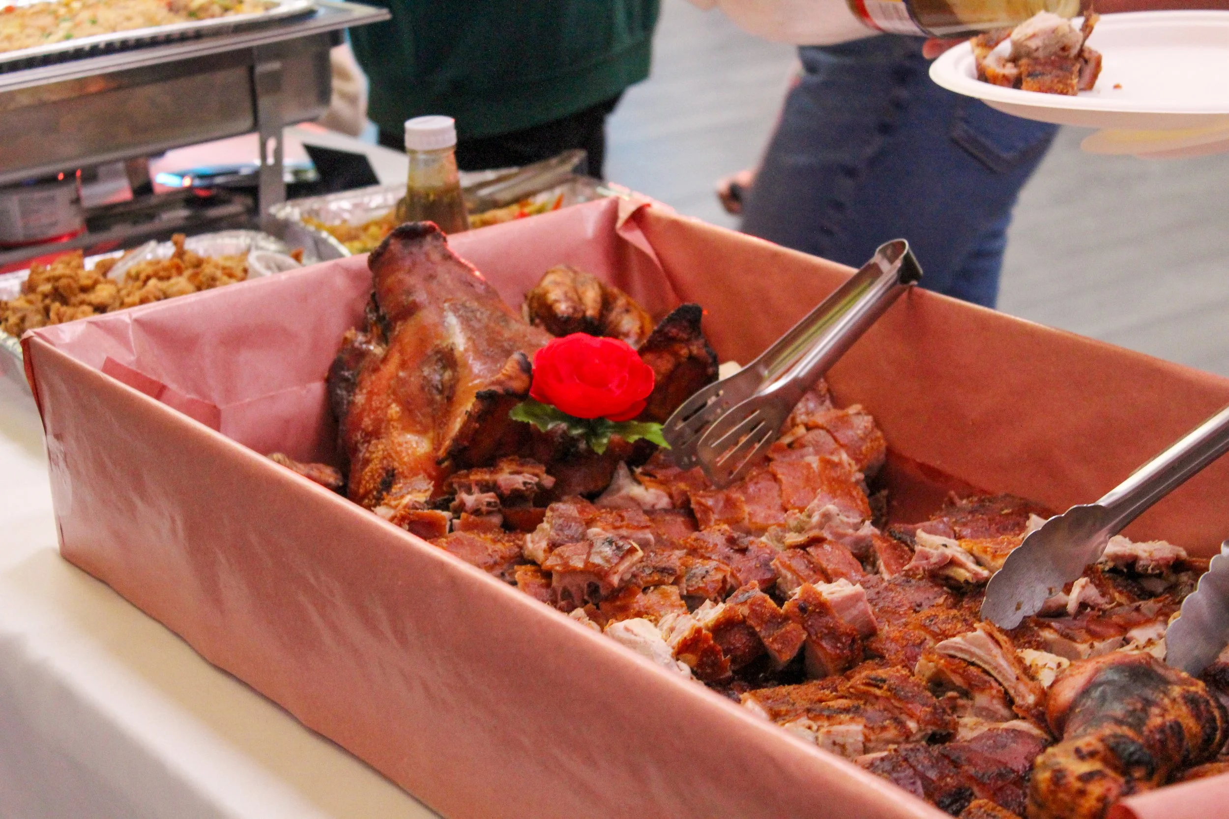A tray of cooked barbecue meats, including a pig's head, with a decorative red flower on top, at a buffet table.