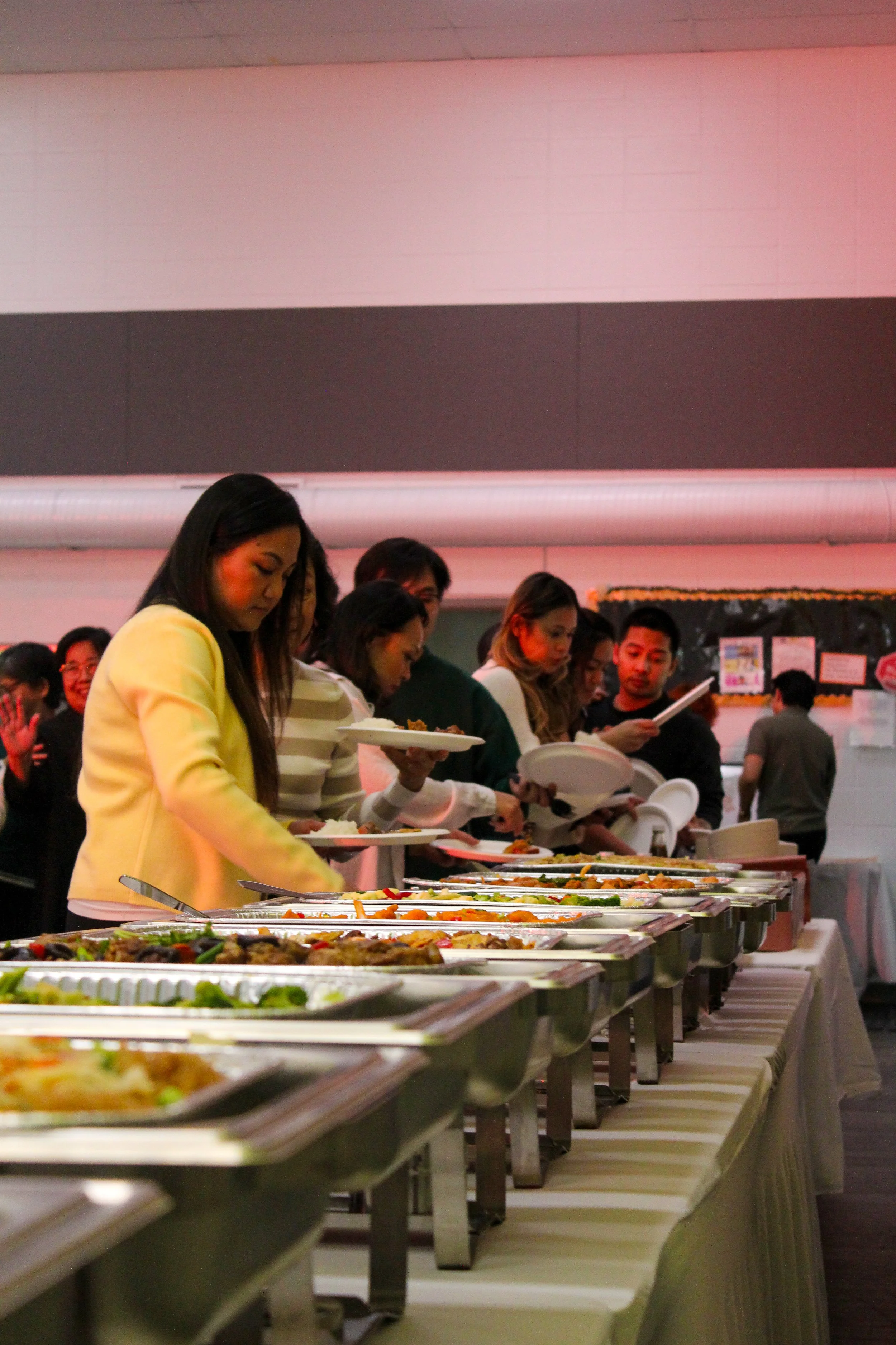 People serving themselves food at a buffet with trays of various dishes on a long table.