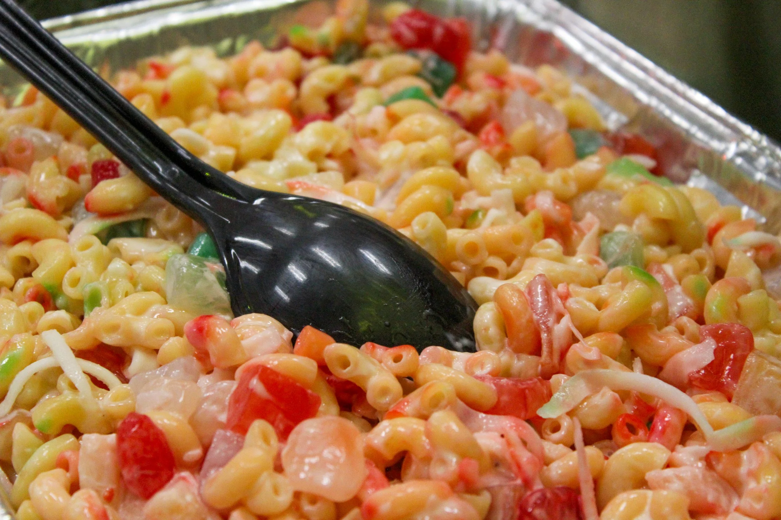 A close-up of a tray of macaroni salad with chopped vegetables, mixed with mayonnaise and served with a black serving spoon.