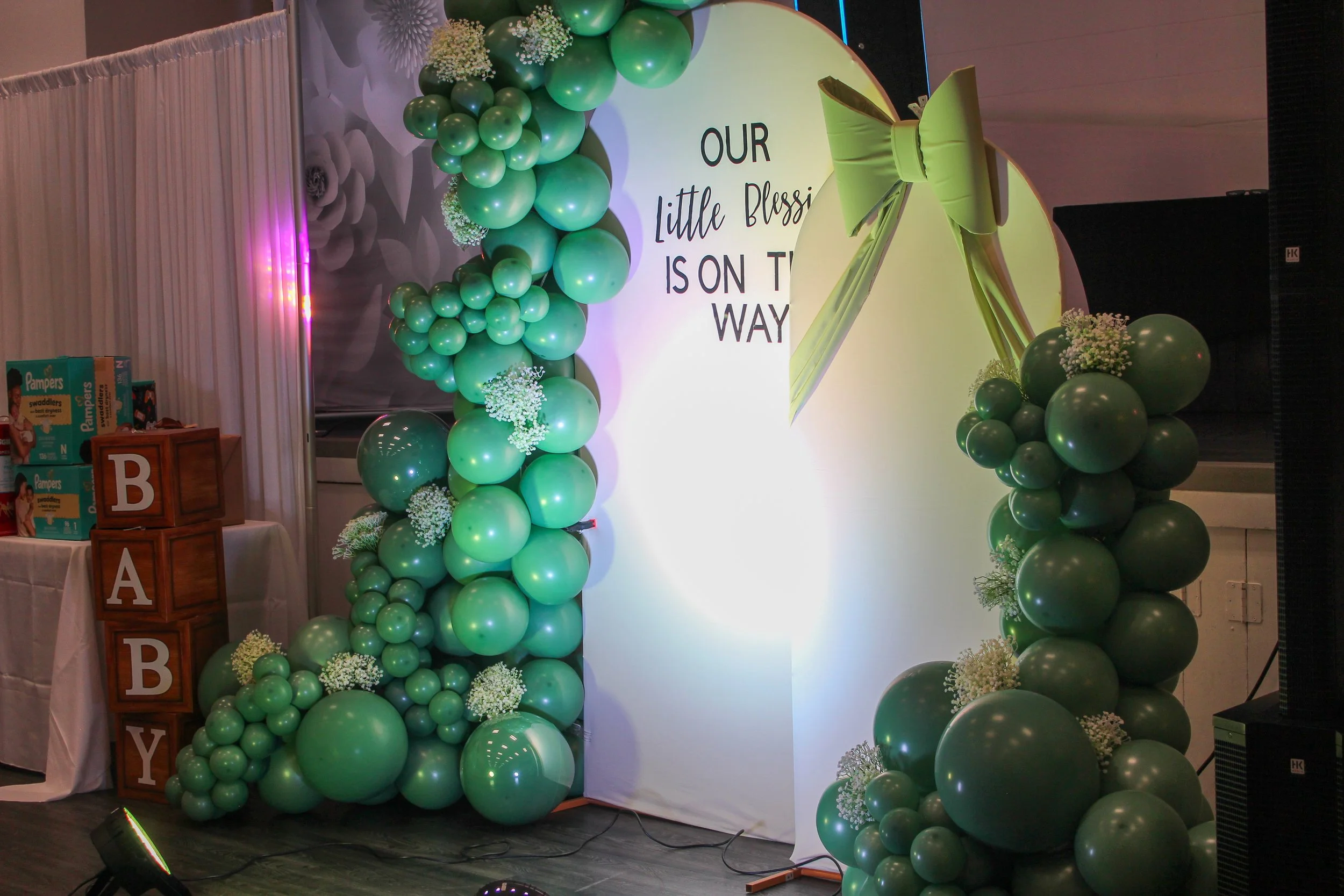 Decorative balloon arch with green balloons and white baby's breath flowers surrounding a white circular backdrop with the wording 'Our Little Blessing is on the Way' and a large green bow.
