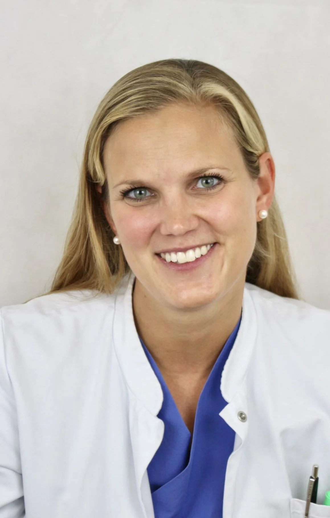 Lächelnde Frau in medizinischer Uniform, vermutlich Ärztin oder Krankenschwester, vor weißem Hintergrund.