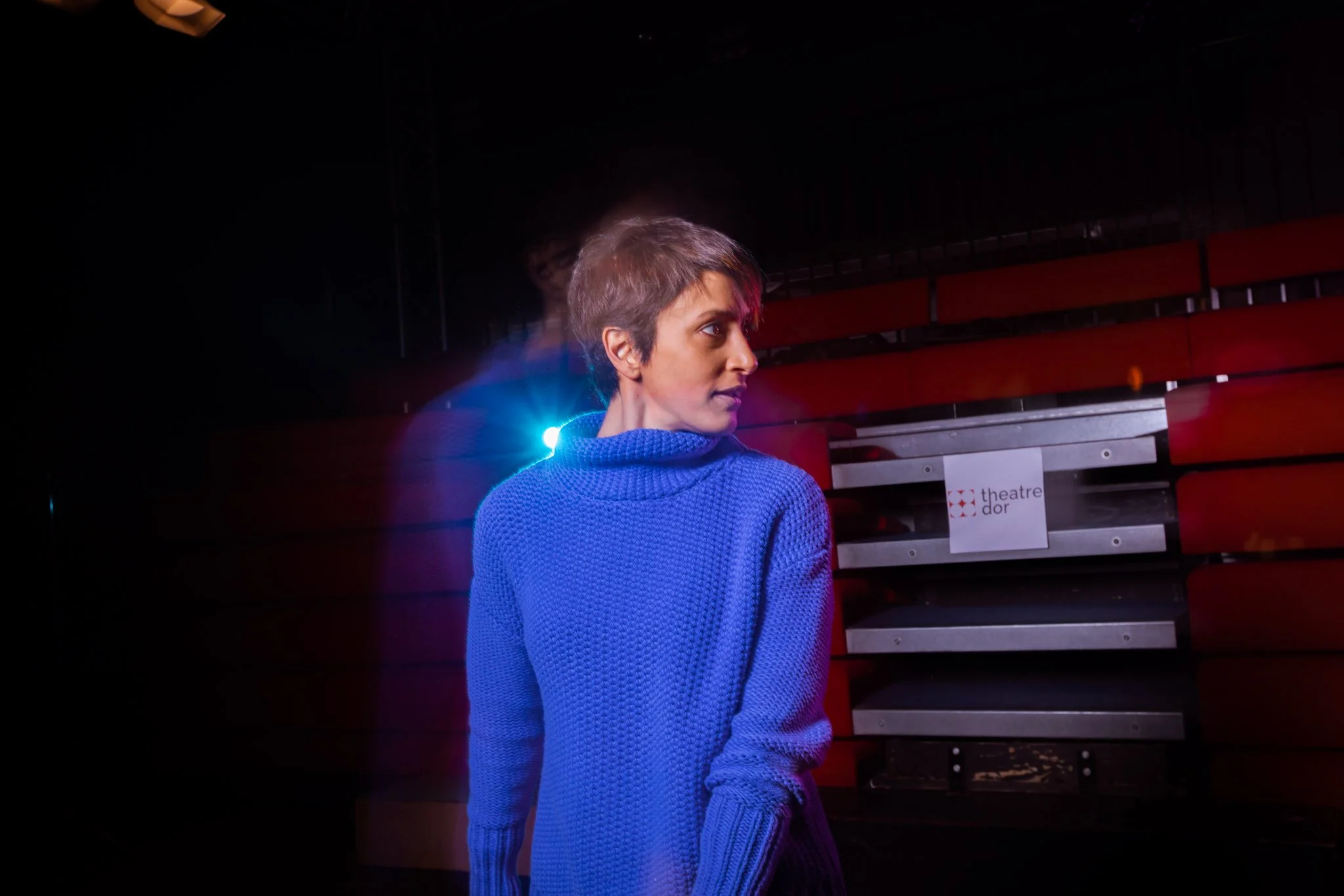 Person in a blue sweater standing in a theater with red seats and blurred lighting.