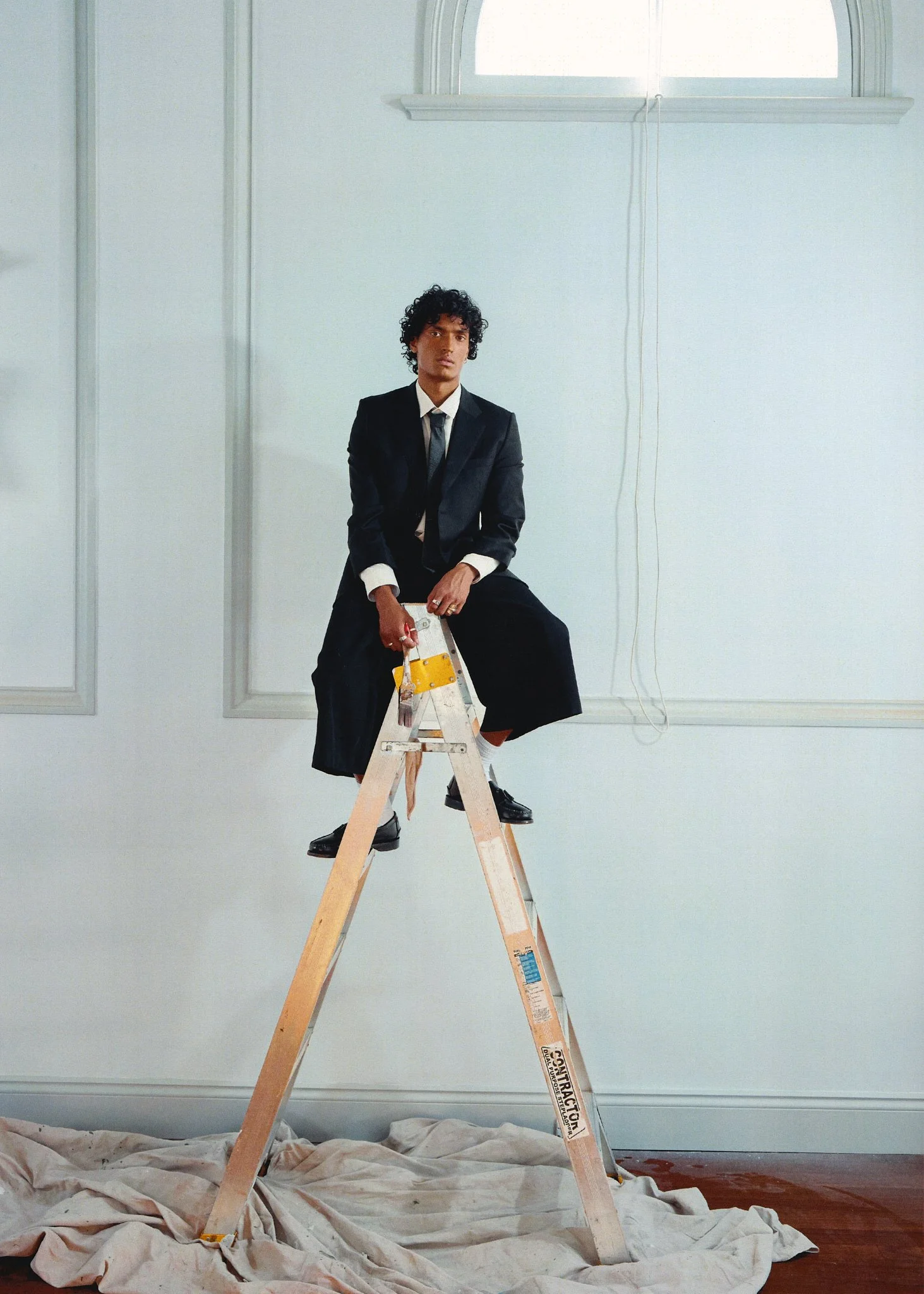 A person in a black suit sitting on a wooden ladder in a bright, white room with high ceilings and decorative wall paneling.