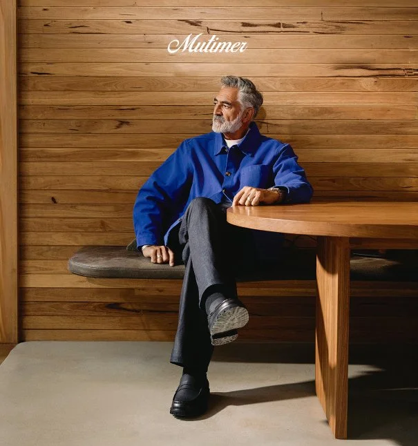 An older man with gray hair and a beard sitting on a booth bench in a restaurant or cafe with wooden walls and furniture, wearing a blue jacket, black pants, and black shoes, looking to his left.