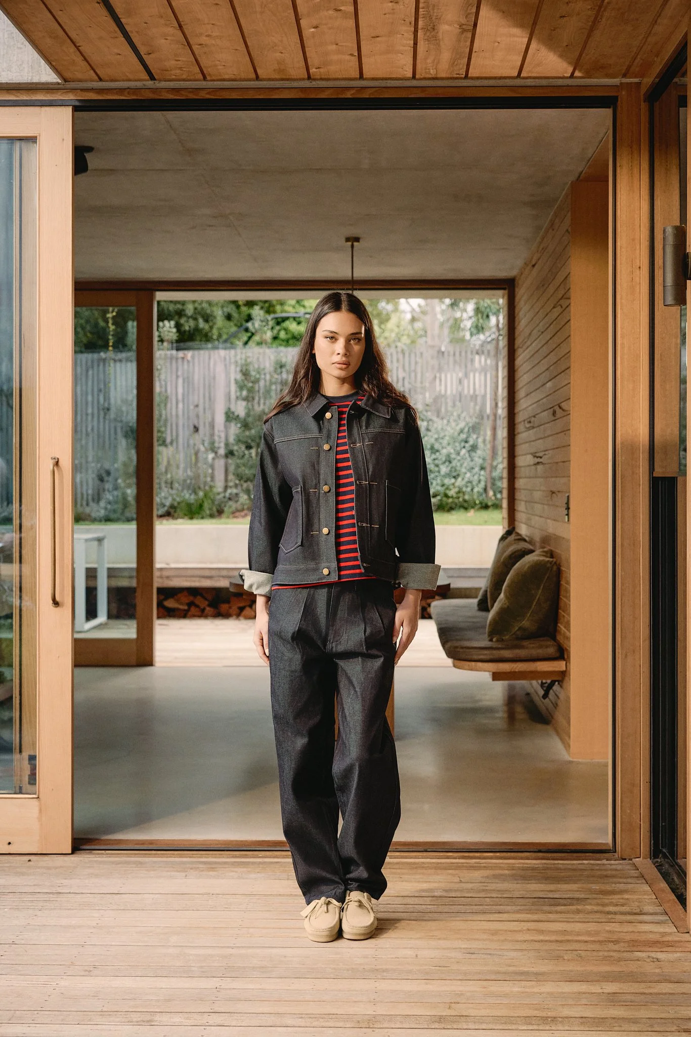 A young woman with long dark hair in a black denim jacket with rolled-up sleeves, black wide-leg trousers, and beige shoes standing in a modern wooden house with a view of a backyard.