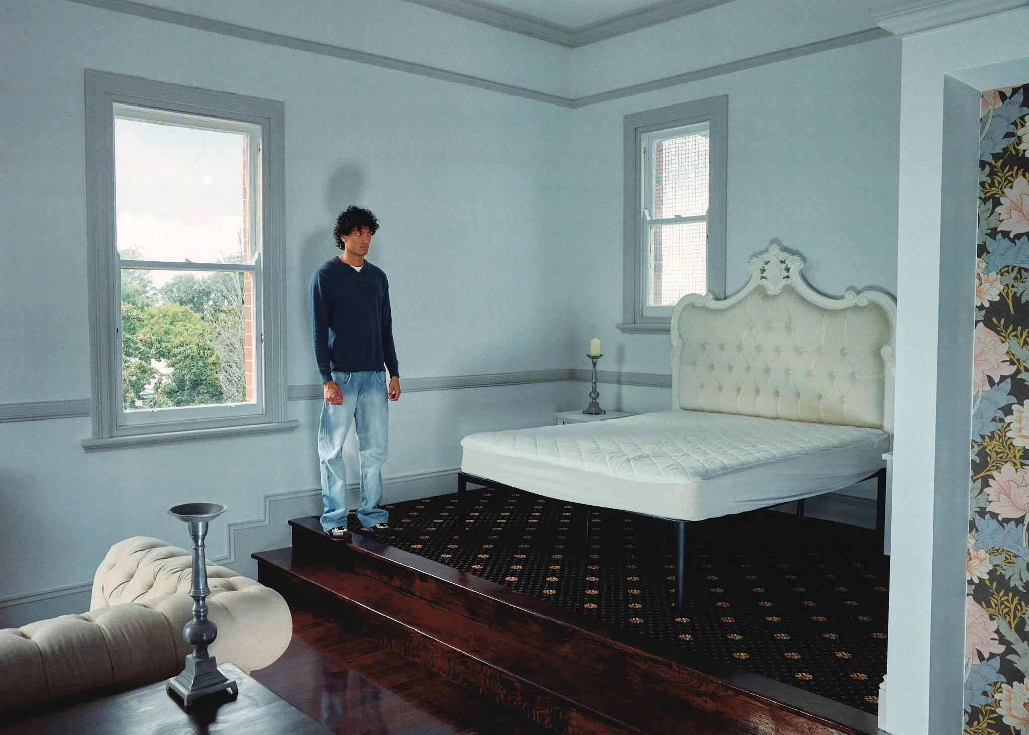 A young man with curly black hair, wearing a dark blue hoodie and light blue jeans, standing on a step inside a bedroom with light subway tiles and a vintage bed with an ornate headboard.