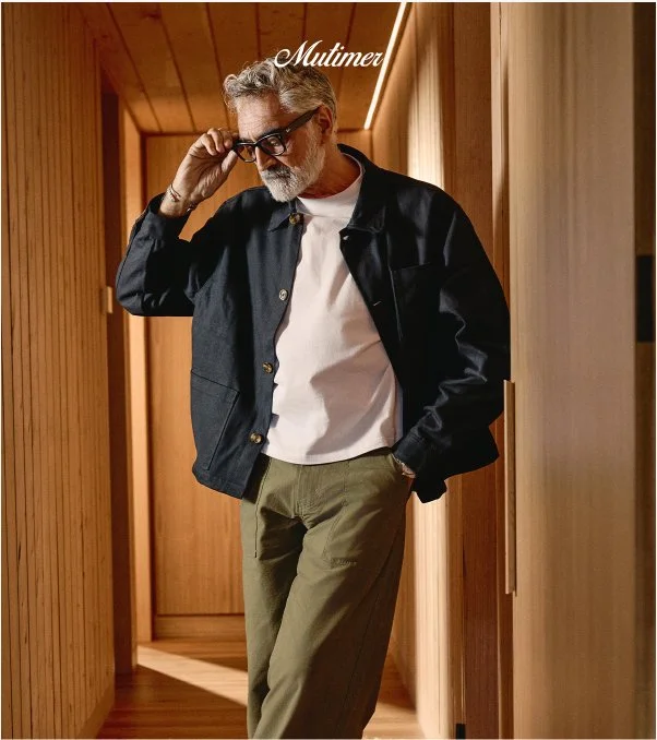 An older man with gray hair and beard standing in a narrow wooden hallway, adjusting his glasses with one hand, wearing a black jacket over a white t-shirt and olive green pants. The word 'Multimer' is written at the top of the image.