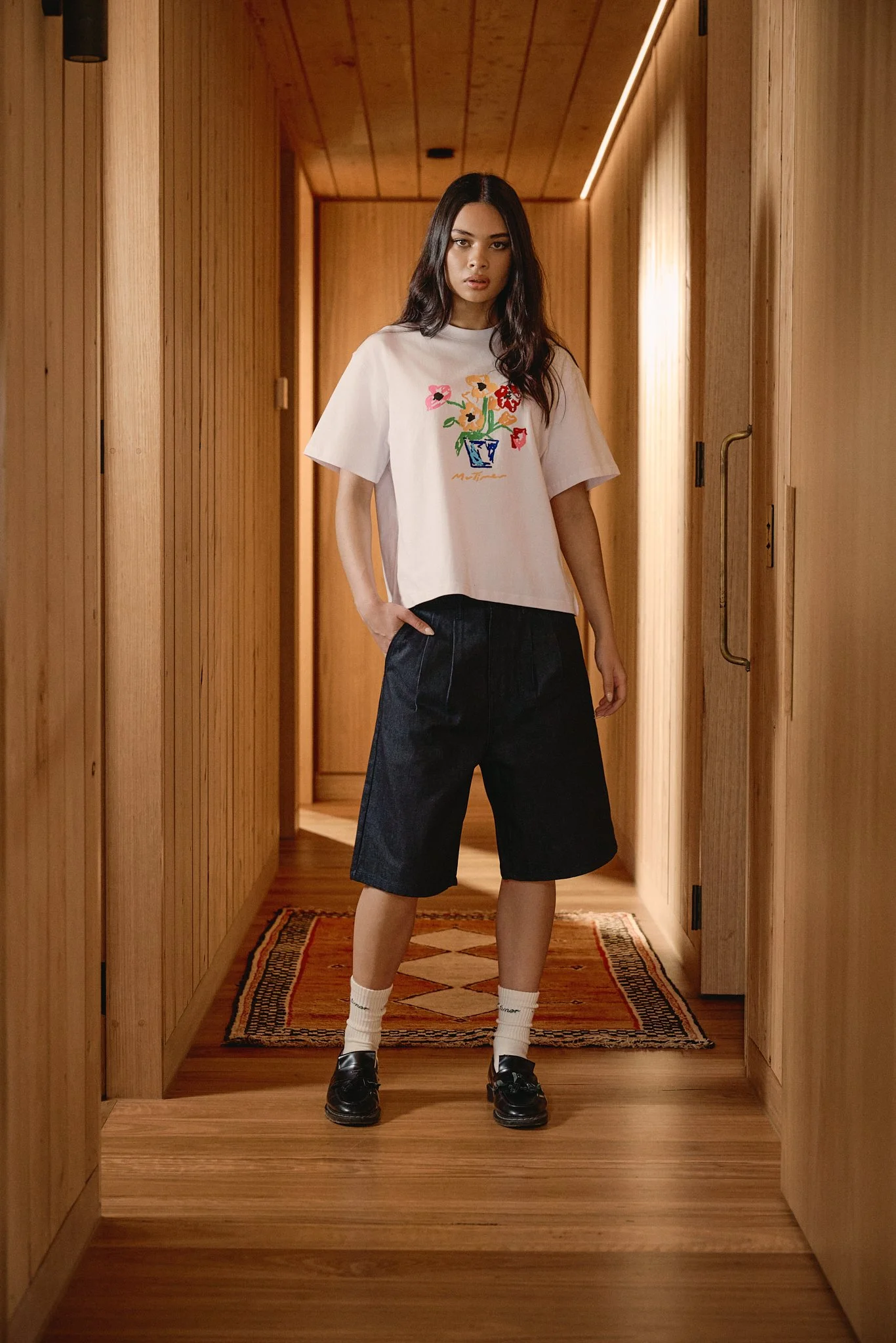 A young woman with long dark hair wearing a white T-shirt with floral embroidery, black knee-length shorts, white socks, and black shoes standing in a wooden hallway.