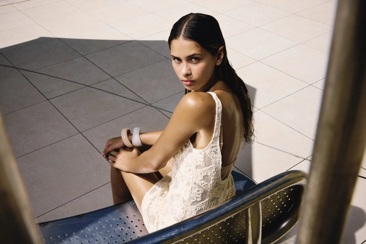 Woman sitting on a bench with her arms crossed, looking up at the camera, wearing a white lace dress and bangles, with a shadow cast behind her on a tiled floor.