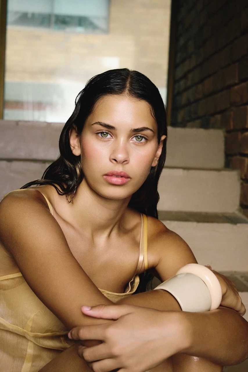 A young woman with dark hair and green eyes sitting on stairs outdoors, wearing a yellow dress and a light-colored bracelet, with a serious expression.