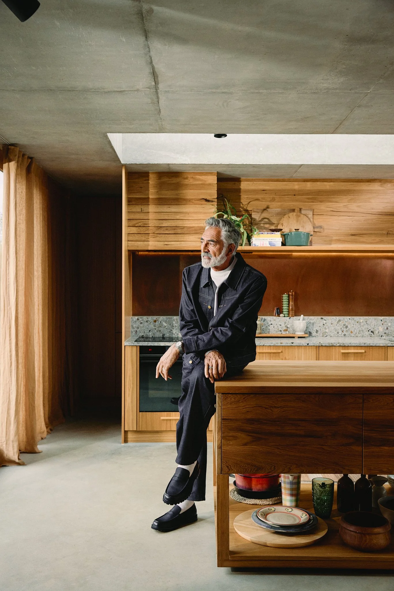 An older man with gray hair and beard, wearing a black jacket and pants, sits on a kitchen island with one leg crossed over the other, in a modern kitchen with wooden cabinets and a granite countertop.