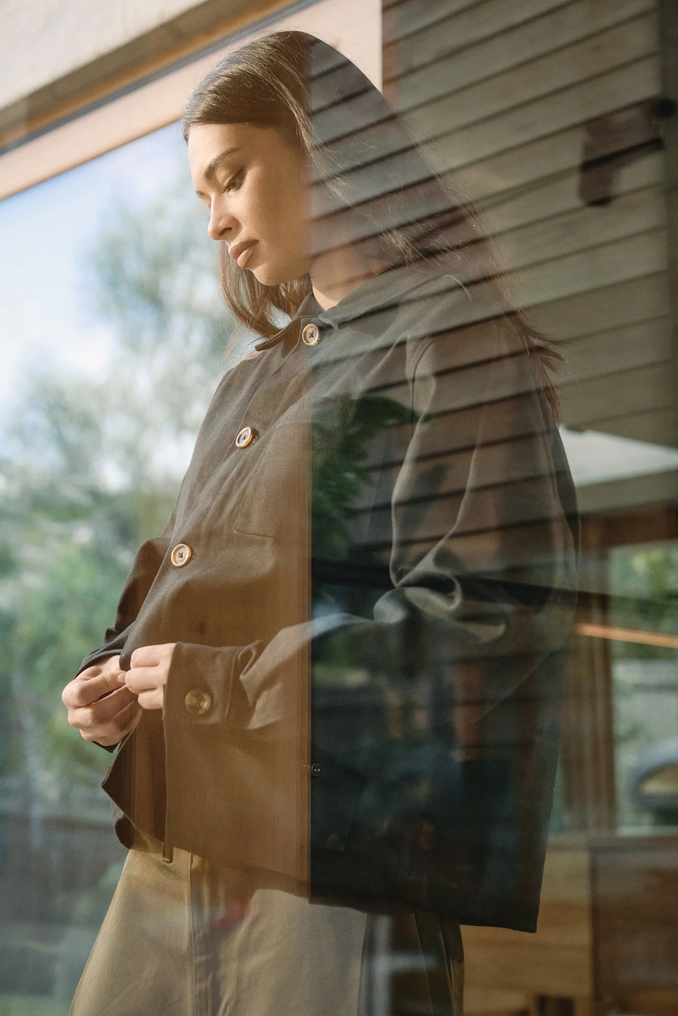 A woman standing inside, seen through a window, adjusting her brown jacket with a contemplative expression, with trees and outdoors visible outside.