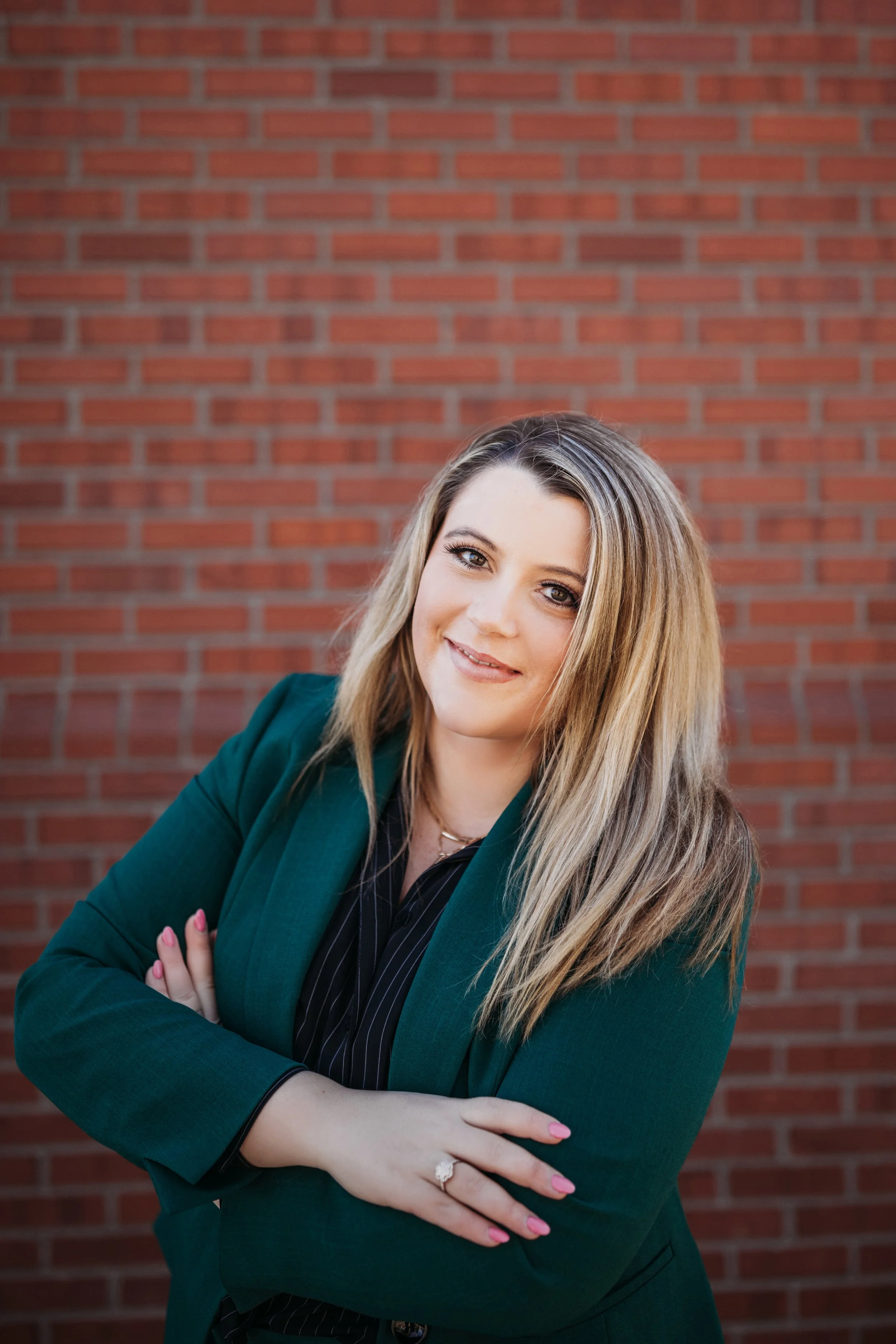 Woman with blonde hair wearing a green blazer standing in front of a brick wall, arms crossed, smiling.