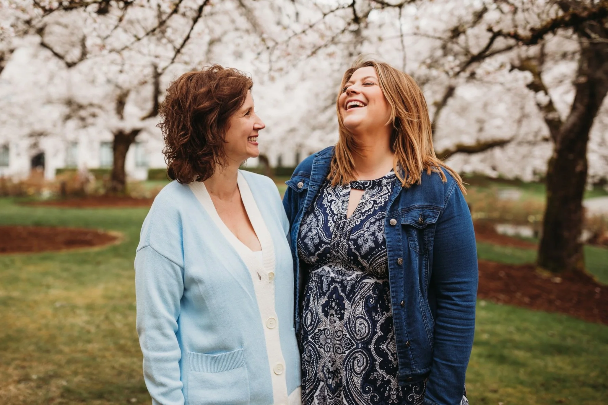 Two women smiling and laughing together outdoors, standing in a park with blooming trees in the background. One woman wears a blue cardigan and the other a blue patterned dress with a denim jacket.