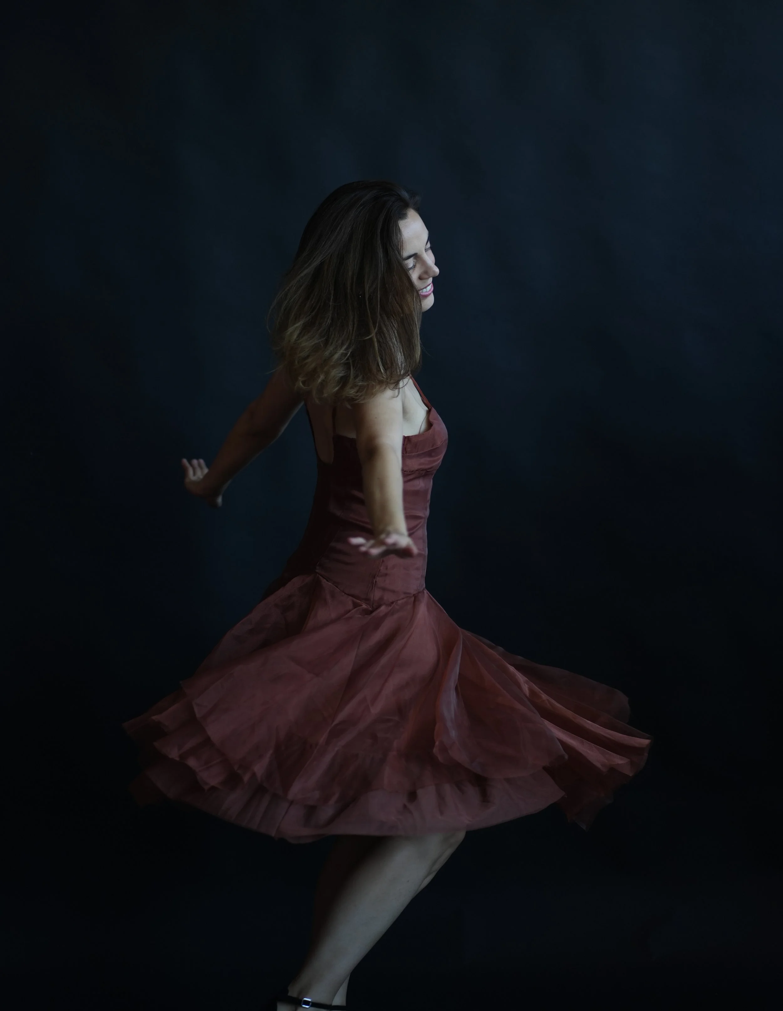 A woman in a brown dress joyfully spinning against a dark background.