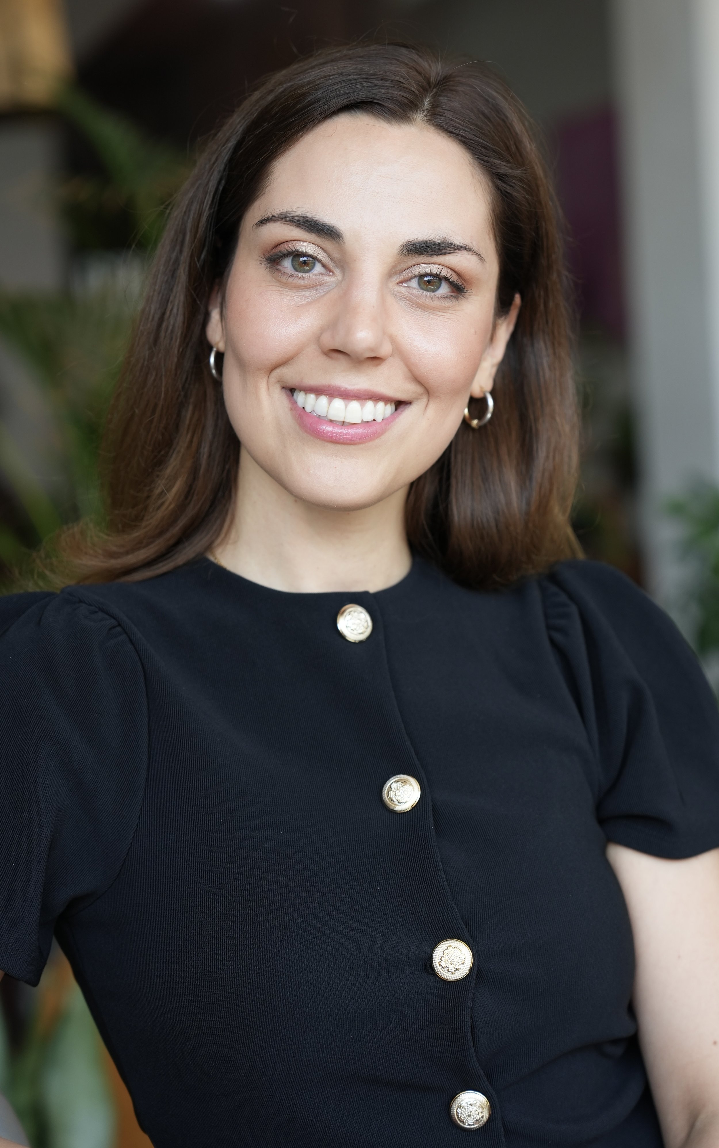 A woman with brown hair, light skin, and blue eyes, smiling, wearing a black top with gold buttons and hoop earrings, indoors with greenery in the background.