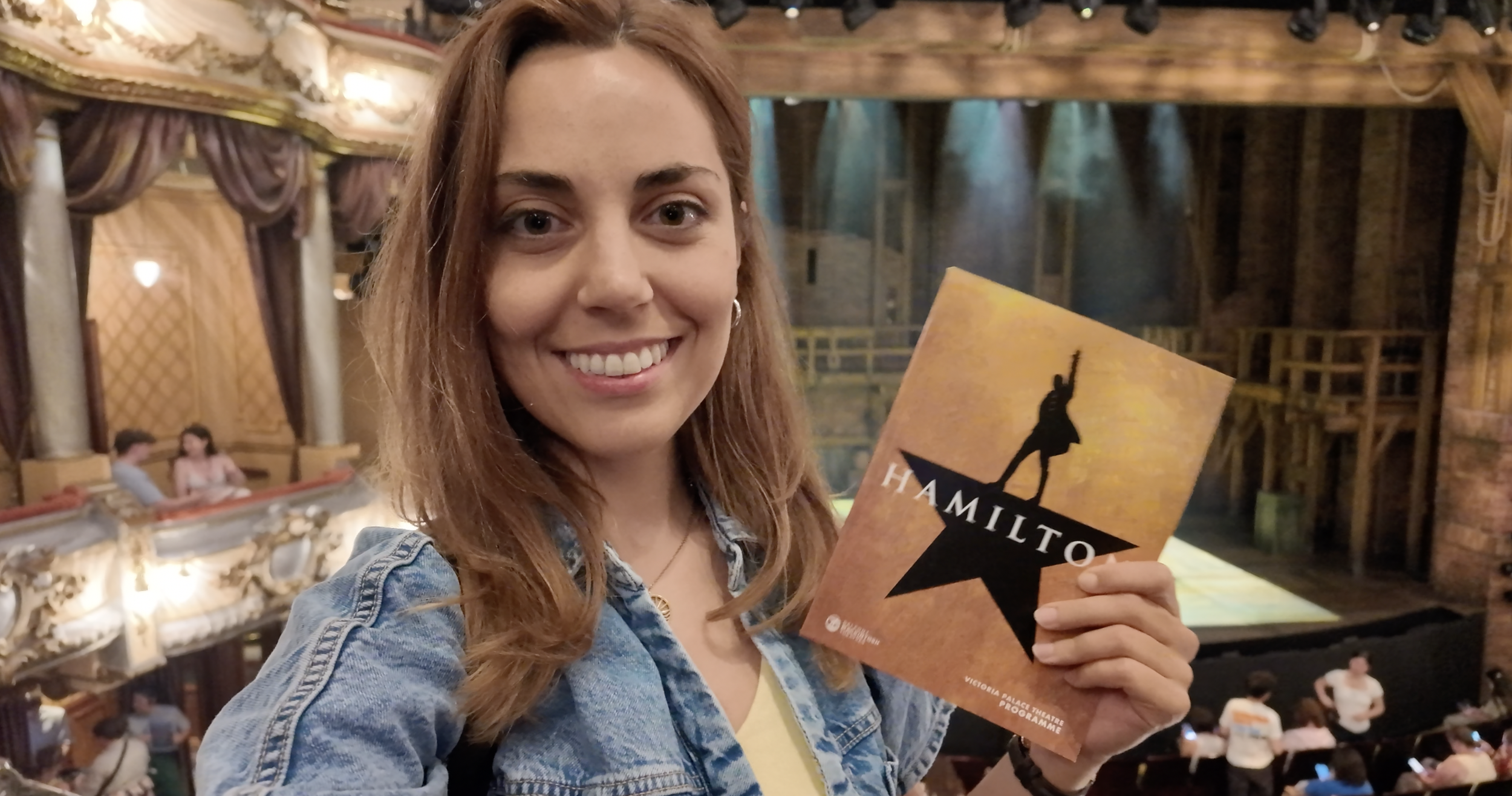 A smiling woman holding a Hamilton theater program in a historic theater with ornate gold and wood detailing, and people seated in the background.