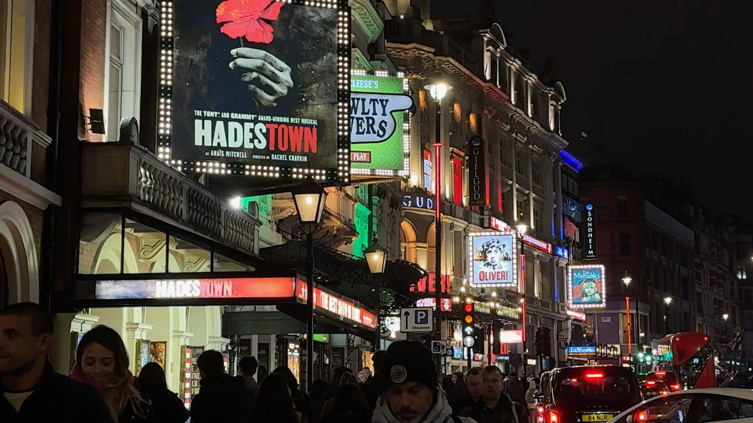 Nighttime street scene with illuminated signs, including a billboard for the musical Hadestown, with a crowded sidewalk and vehicles on the street.